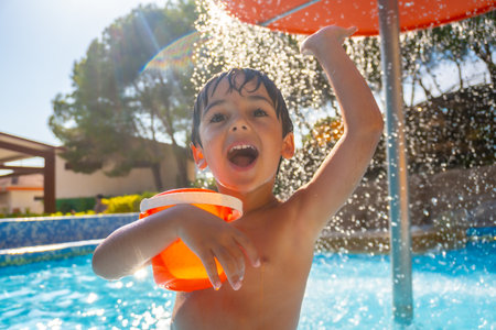 Boy holding orange bucket and raising arm under refreshing water shower in swimming pool on sunny summer dayの写真素材