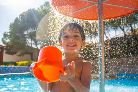Smiling child enjoying a sunny day at the pool, holding an orange bucket under a refreshing water fountain, surrounded by trees and clear skyの写真素材