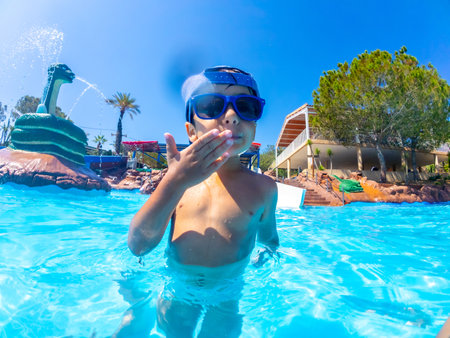 Young boy wearing sunglasses blowing a kiss while standing in a swimming pool at a water park with a snake shaped fountain in the backgroundの写真素材