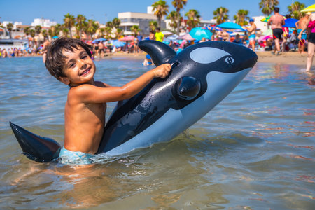 Smiling child having fun riding an inflatable orca toy in the shallow sea water by the beach on a sunny summer dayの写真素材