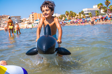 Smiling boy riding an inflatable orca toy in the sea waves, enjoying summer vacation at a crowded beachの写真素材