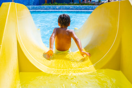 Boy joyfully sliding down a colorful waterslide in an aquapark, enjoying summer vacation with laughter and excitement under the sunny skyの写真素材