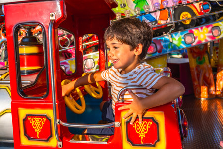 Young boy smiling while steering a vibrant red amusement park ride, surrounded by colorful decorations and lights, capturing the essence of childhood funの写真素材