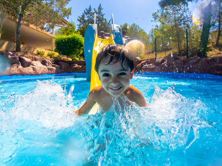 Young boy enjoying a sunny day, sliding into a vibrant blue pool with a big splash, surrounded by trees and clear skiesの写真素材