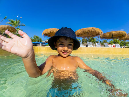 Smiling child in a sun hat waves while playing in a clear swimming pool, surrounded by straw umbrellas and tropical palm treesの写真素材