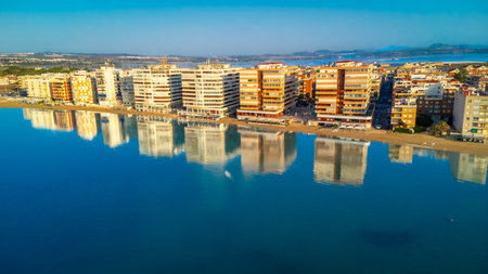 Aerial view of Torrevieja skyline reflecting on calm Mediterranean sea at sunrise, with colorful buildings and tranquil waters creating a peaceful sceneの写真素材