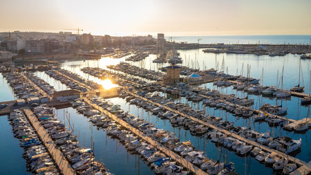 Golden sunrise reflecting on calm waters of Torrevieja marina, showcasing docked boats and the cityscape in the backgroundの写真素材