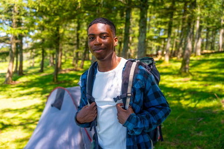 Young black man wearing a backpack, enjoying a camping trip in the woods, surrounded by lush trees...