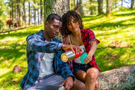Young couple sharing a hot drink from a thermos while taking a break during their hike in a sunny forestの写真素材