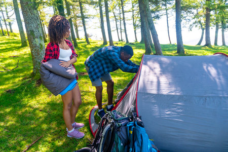 Couple setting up a tent in a lush forest, preparing their campsite for a summer adventure filled...