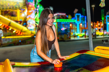 Happy woman playing air hockey at amusement park, enjoying nightlife entertainment with colorful lights in backgroundの写真素材