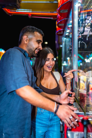 Young couple having fun playing a claw crane game at an amusement park at night, trying to win a prizeの写真素材
