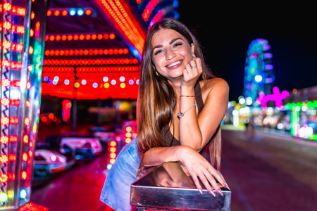 Happy woman enjoying night at amusement park, leaning on railing with colorful bumper cars and ferris wheel in backgroundの写真素材