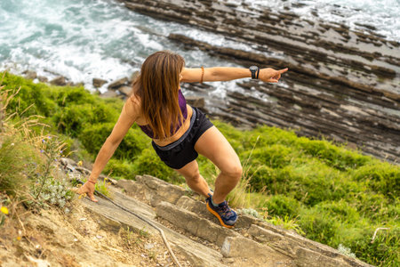 Female athlete descending steep stone stairs and pointing at the geological formations of the Basque...