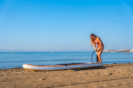 Woman inflating a paddleboard on a sandy beach, ready for a summer day of paddle surfing under a...