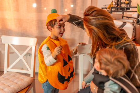 Mother applying halloween makeup on her son wearing a pumpkin costume, while holding her babyの写真素材