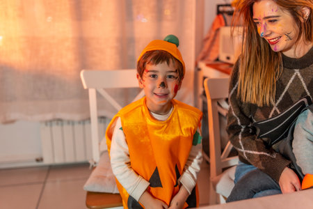 Happy child wearing pumpkin costume smiling and looking away while sitting next to his mother during halloween party at homeの写真素材
