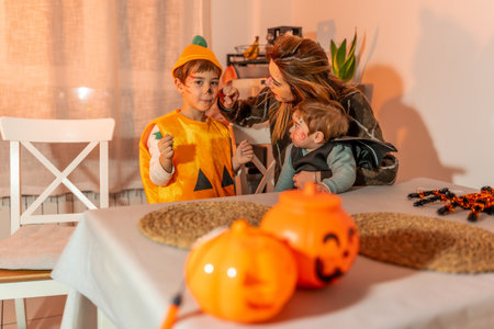 Mother applying halloween makeup on her children, a boy dressed as a pumpkin and a baby in a bat costumeの写真素材