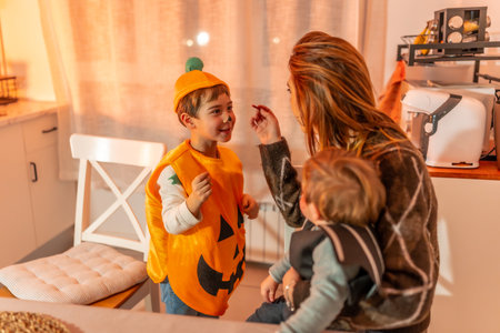 Mother applying halloween makeup to her son wearing a pumpkin costume while holding her baby in the kitchenの写真素材