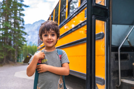 Happy elementary school student carrying backpack is waiting for the school bus in a mountain location, ready for his first day back to schoolの写真素材