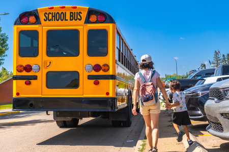 Mother holding son's hand walking towards a school bus on the first day back to school, representing...