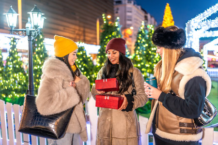 Three friends exchanging Christmas presents and enjoying hot drinks at a decorated outdoor Christmas market in a city at nightの写真素材