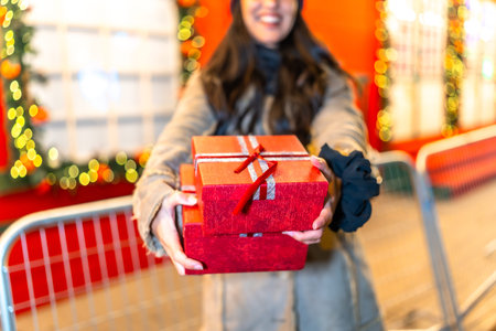 Woman holding Christmas presents at a festive market, smiling brightly while sharing the joy of...