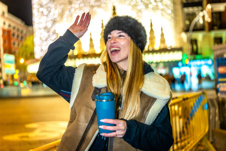 Young woman greeting someone while enjoying a hot beverage in a reusable cup in a city decorated...