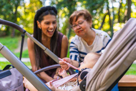 Grandmother feeds baby girl sitting in stroller, while mother watches, enjoying family time together in parkの写真素材