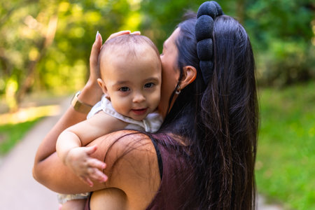 Mother cuddling her baby girl while strolling through a park, savoring a sunny day filled with family love and joyful bondingの写真素材