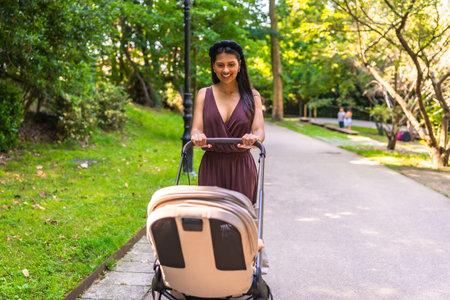 Latin mother pushing a stroller with a happy baby inside, enjoying a sunny day while walking through a peaceful park filled with greeneryの写真素材