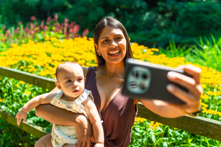 Happy latin mother holding her baby daughter and taking a selfie with smartphone in a beautiful garden full of yellow flowersの写真素材