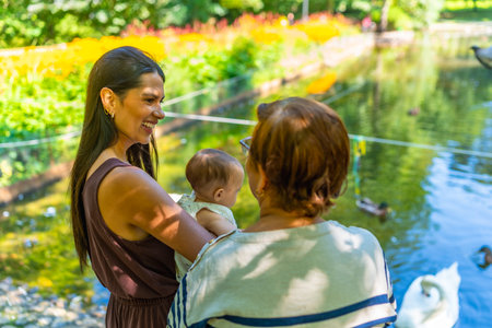 Three generations of a latin family, grandmother, mother, and baby granddaughter, spending quality time together at a park, enjoying the view of a pond with swansの写真素材