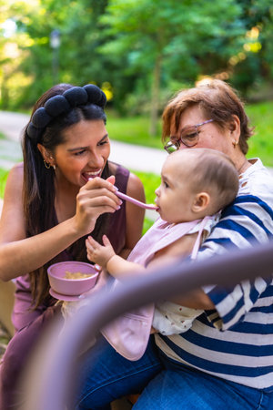 Mother feeding her baby a meal in the park, with grandmother holding the child, enjoying family time togetherの写真素材
