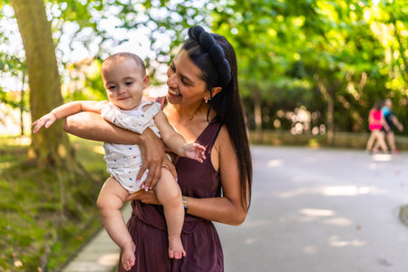 Happy latin mother holding and playing with her baby daughter in a park, enjoying a sunny day outdoorsの写真素材