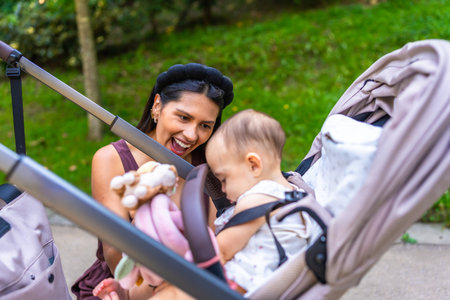 Happy latin mother showing toys to her baby daughter sitting in a stroller, enjoying a sunny day together in a parkの写真素材