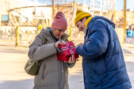 Happy senior couple sharing a joyful moment while opening a festive Christmas gift box in a charming winter city settingの写真素材