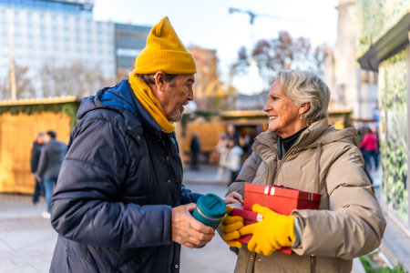 Happy senior couple exchanging Christmas gifts and enjoying a warm beverage at a bustling outdoor...