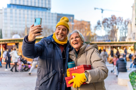 Cheerful senior couple taking a selfie with smartphone at a Christmas market, holding gifts and enjoying winter holidaysの写真素材