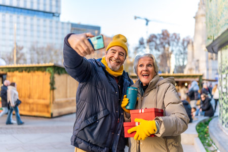 Elderly couple taking a selfie with smartphone at a Christmas market, holding gifts and hot drinks, enjoying winter holidays in the cityの写真素材