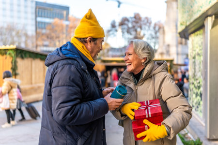 Smiling elderly couple exchanging Christmas presents and enjoying hot beverages at a winter market, celebrating the festive season togetherの写真素材