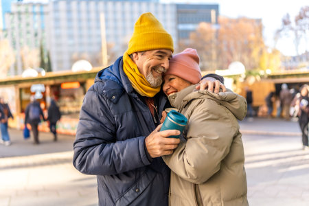 Elderly couple hugging and smiling at an outdoor Christmas market, enjoying a warm beverage on a cold winter dayの写真素材