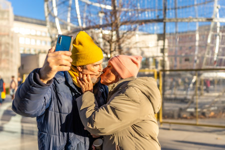 Happy senior couple kissing and taking a selfie with smartphone in a winter city during christmas...
