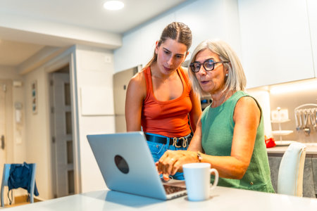Senior woman navigating a laptop with help from a younger woman in a cozy kitchen, fostering family connection and digital skillsの写真素材