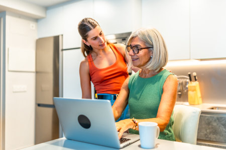 Senior woman is using a laptop in a kitchen while a young woman stands behind her, providing assistance and supportの写真素材