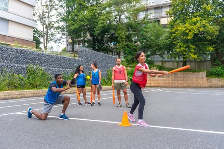 Group of friends enjoying a lively game of baseball on an outdoor court, with a woman swinging an...