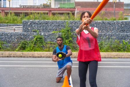 Engaging in an outdoor baseball practice, a woman swings a bat with focus while a man kneels, poised...