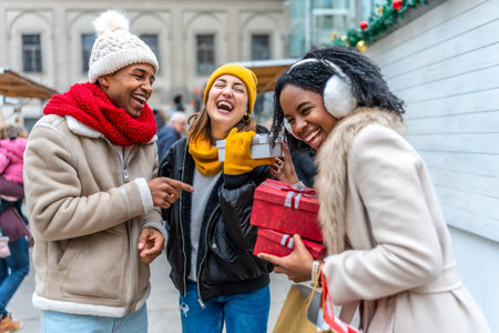 Three friends are having fun exchanging Christmas gifts at a winter market, laughing and enjoying...