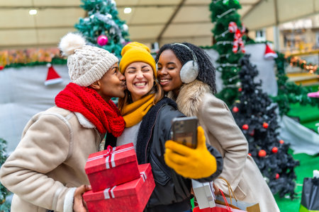 Three happy friends are taking a selfie at a Christmas market, one of them is kissing another one on the cheek while holding Christmas giftsの写真素材