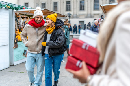Happy multi ethnic couple walking arm in arm between christmas market stalls, woman carrying presents